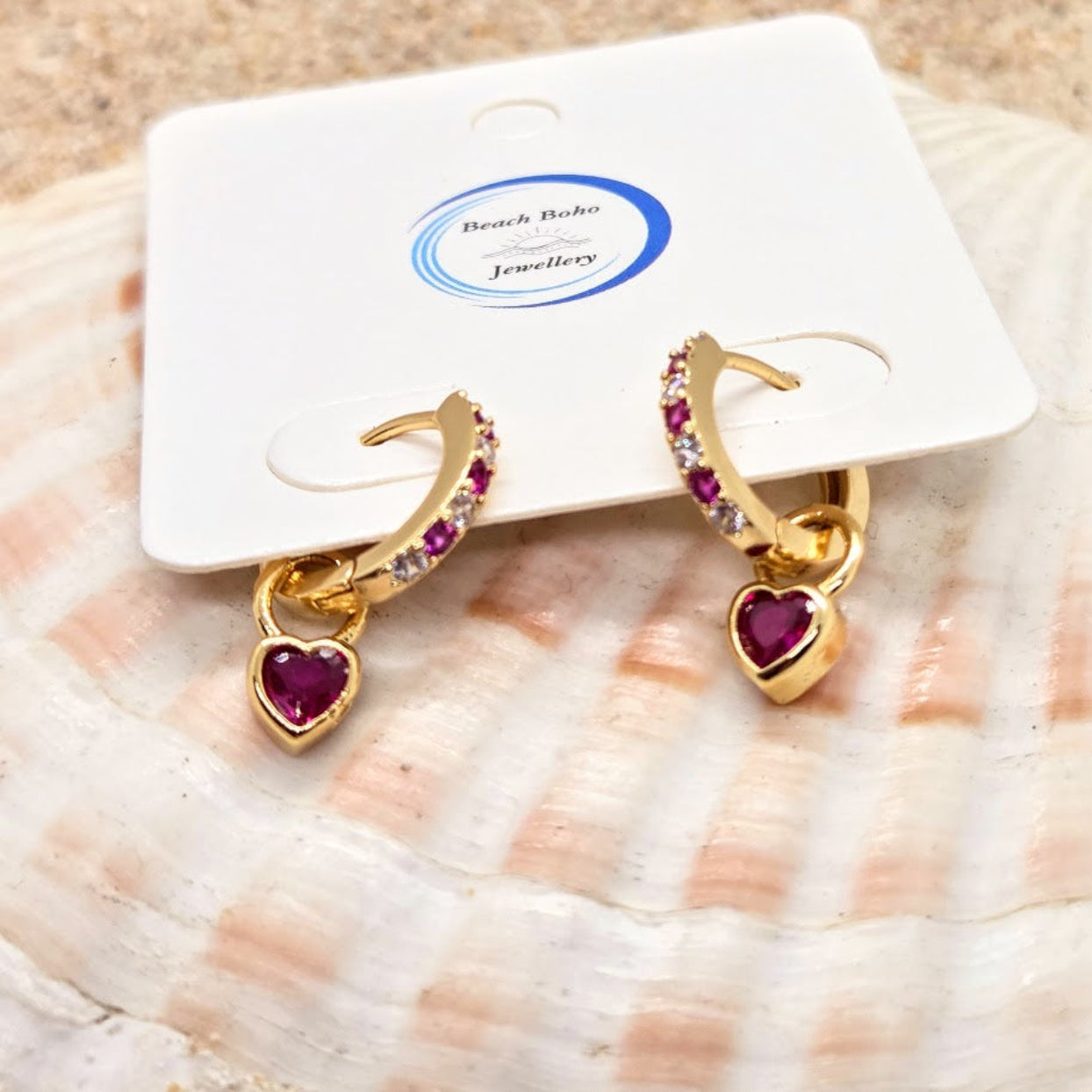 Gold earrings with red heart charms on a white card with 'Beach Babe Jewellery' branding, placed on a seashell.