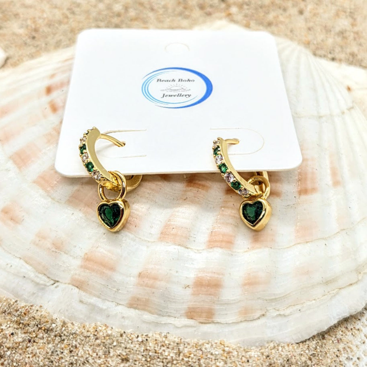 Gold hoop earrings with green heart charms on a shell with 'Beach Babe Jewellery' packaging.