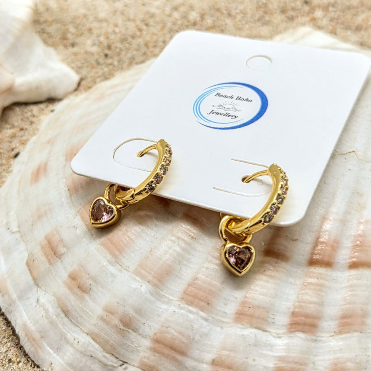 Gold heart-shaped earrings on a white card with 'Beach Bazaar Jewelry' label, placed on a seashell.