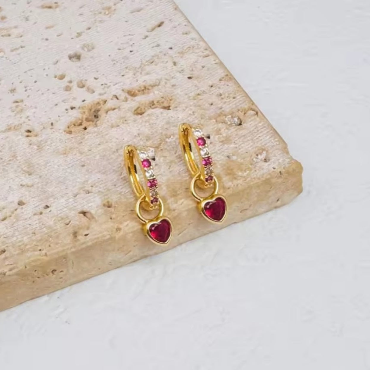 Gold hoop earrings with red heart charms on a stone surface