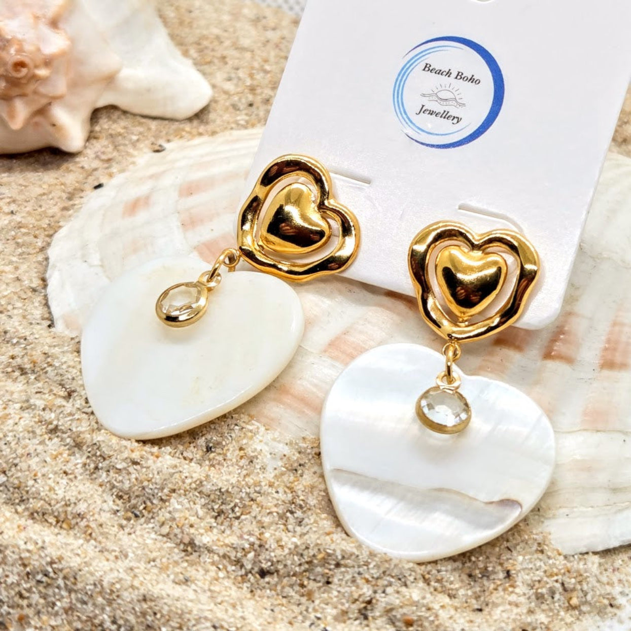 Gold heart-shaped earrings with white stones on a shell with 'Beach Babe Jewellery' card.