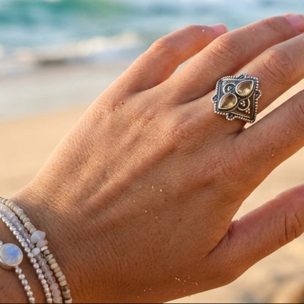 A close-up of a hand wearing a silver-toned ring with a floral or clover-like motif, set against a blurred beach background with ocean waves and rocks.