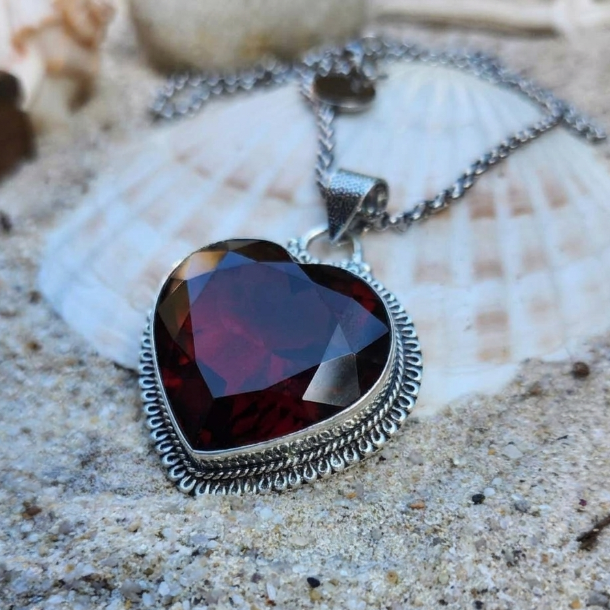 A heart-shaped faceted dark red gemstone pendant in a textured silver bezel setting, resting on a white seashell over sand.