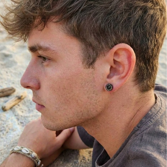 A close-up side profile of a man on a sandy beach wearing a black circular stud earring with a silver-toned center, along with a silver bracelet.