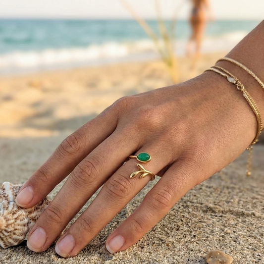 A gold ring with a green oval stone worn on a person's ring finger, shown on a sandy beach with the ocean in the background.