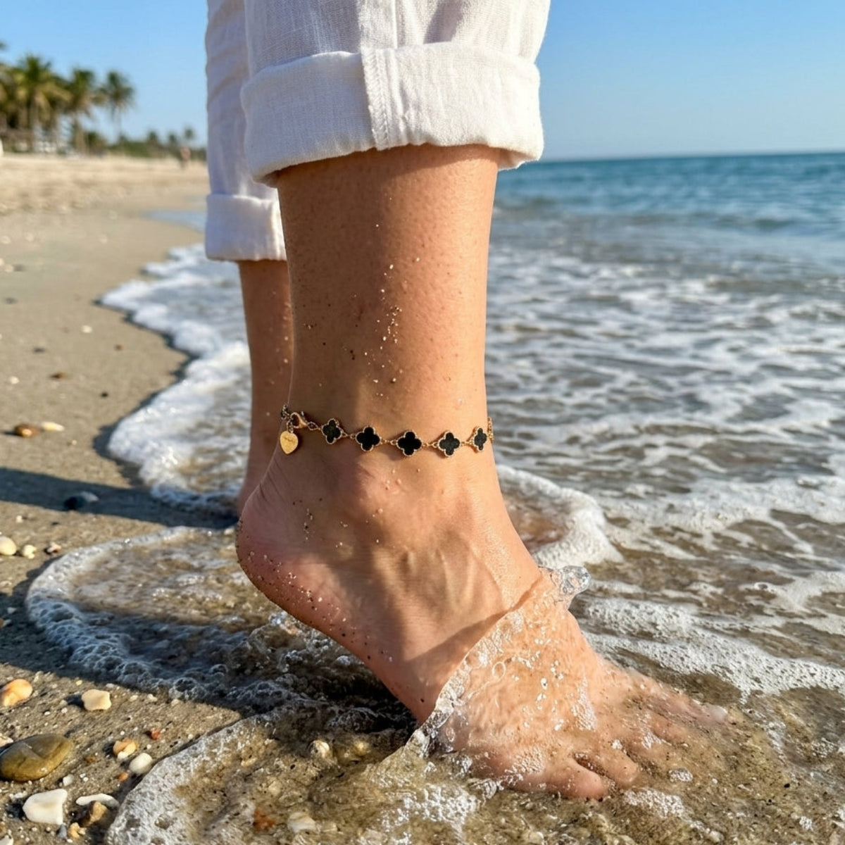 A close-up of a person's bare ankle and foot standing in shallow ocean water on a sandy beach, wearing a delicate black beaded anklet with small gold heart charms.