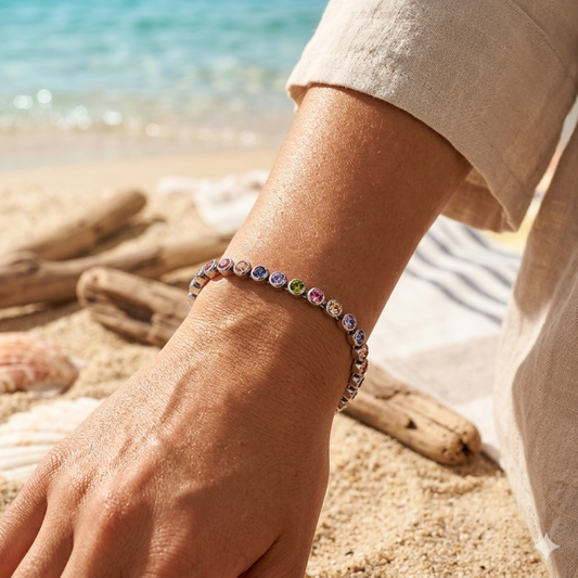 Hand wearing a colorful beaded bracelet on a beach with shells and driftwood.