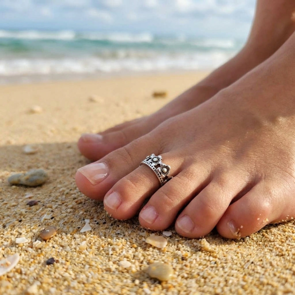 A silver toe ring with a floral crown design worn on a person's toe, set against a sandy beach background with the ocean and sky blurred in the distance.