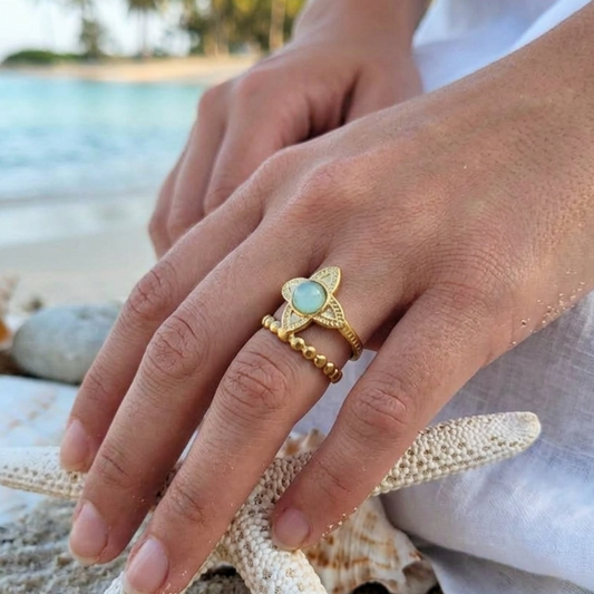 A close-up of a person's hand wearing a gold ring with a light blue-green stone, styled on a beach with starfish and seashells in the background.