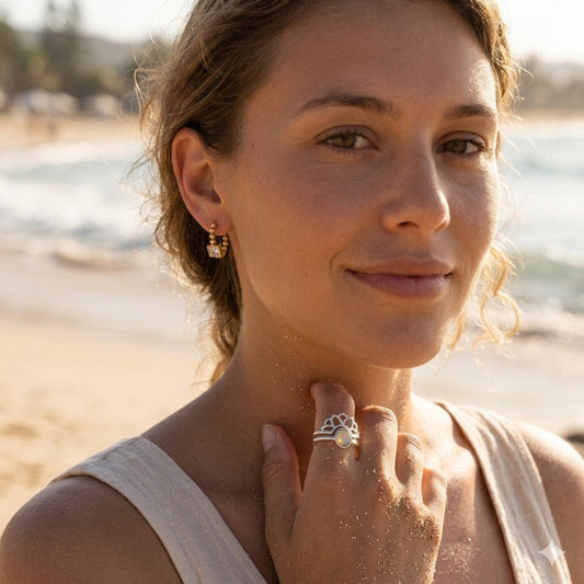 A woman on a sandy beach wearing a silver ring with a white stone and a small gold earring, with the ocean blurred in the background.