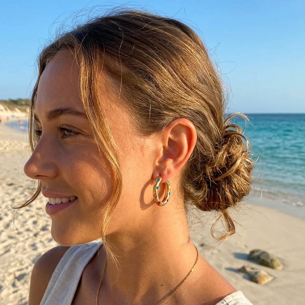 A woman with her hair in a low bun stands on a sandy beach wearing gold hoop earrings and a delicate gold necklace. The background shows the blue ocean and sky.
