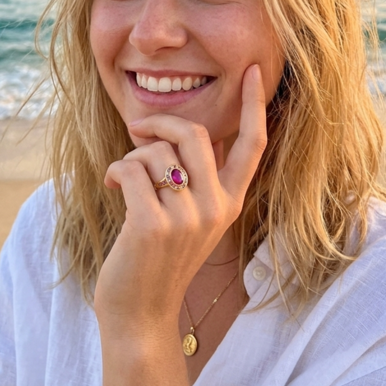 A smiling blonde woman posing on a beach at sunset, wearing a white button-up shirt, a delicate gold necklace, and a gold ring with a pink gemstone.