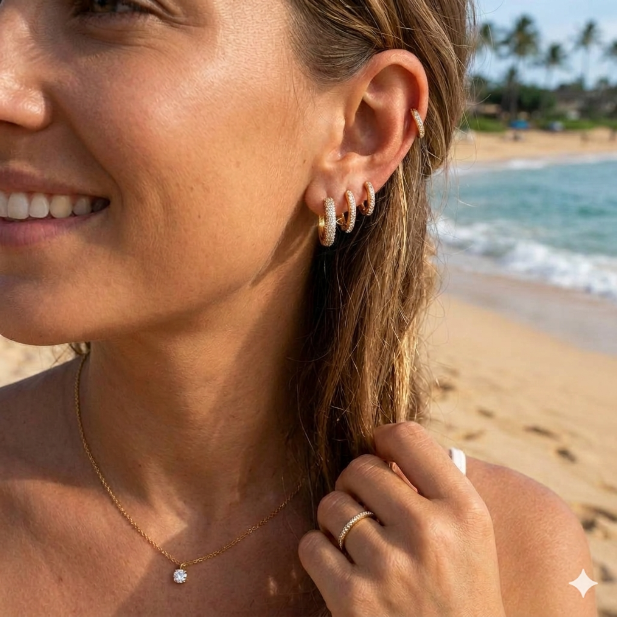 Woman on a beach wearing gold hoop earrings and a necklace, with a blurred background of ocean and sky.