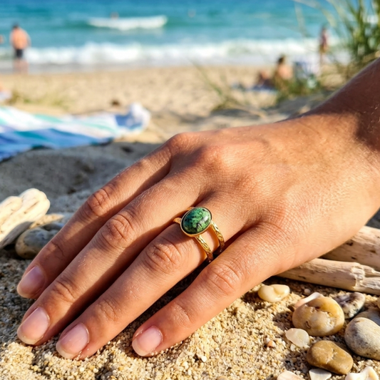 A gold ring with a green oval stone worn on a person's hand, resting on sandy beach next to driftwood and seashells, with the ocean and beachgoers blurred in the background.