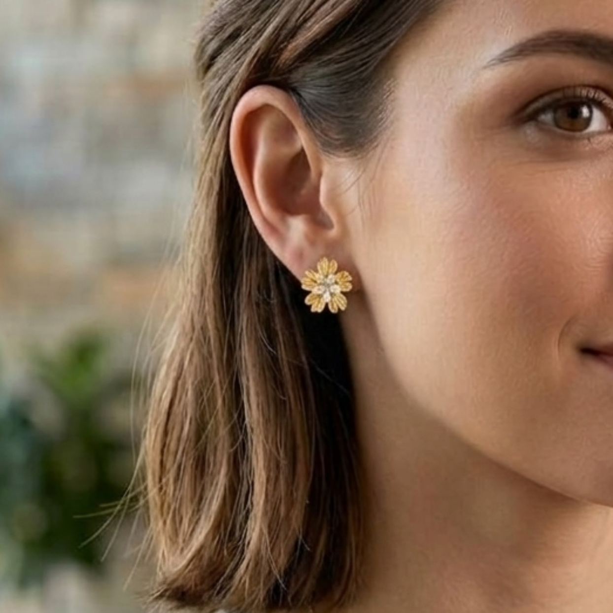 A woman with shoulder-length brown hair wearing a cream ribbed knit sweater and gold jewelry, including a small flower-shaped earring and a delicate necklace, in a softly blurred indoor setting.