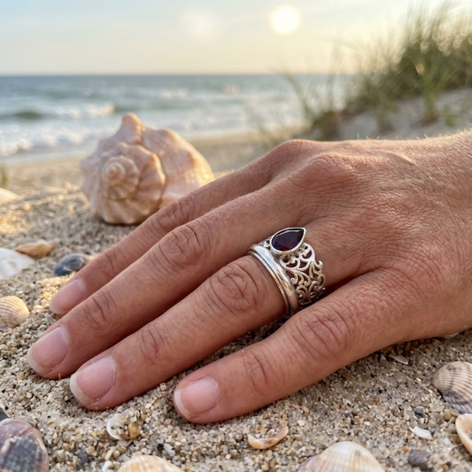 Hand wearing a silver ring with a red stone on a sandy beach with shells and ocean in the background
