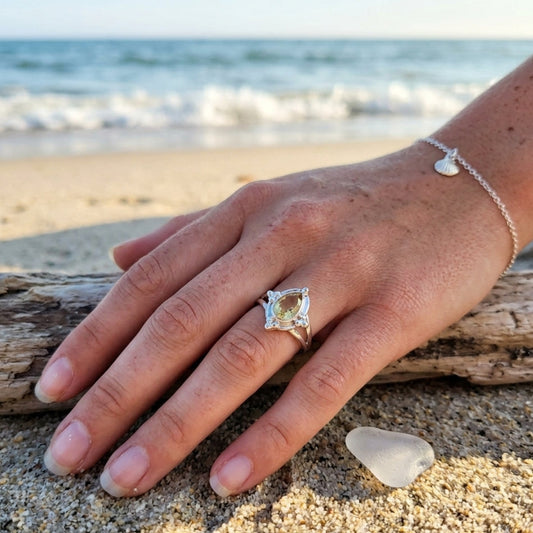 A silver ring with a square-cut pale yellow gemstone is worn on a person's hand resting on a beach, with driftwood and sand in the foreground and the ocean in the background.