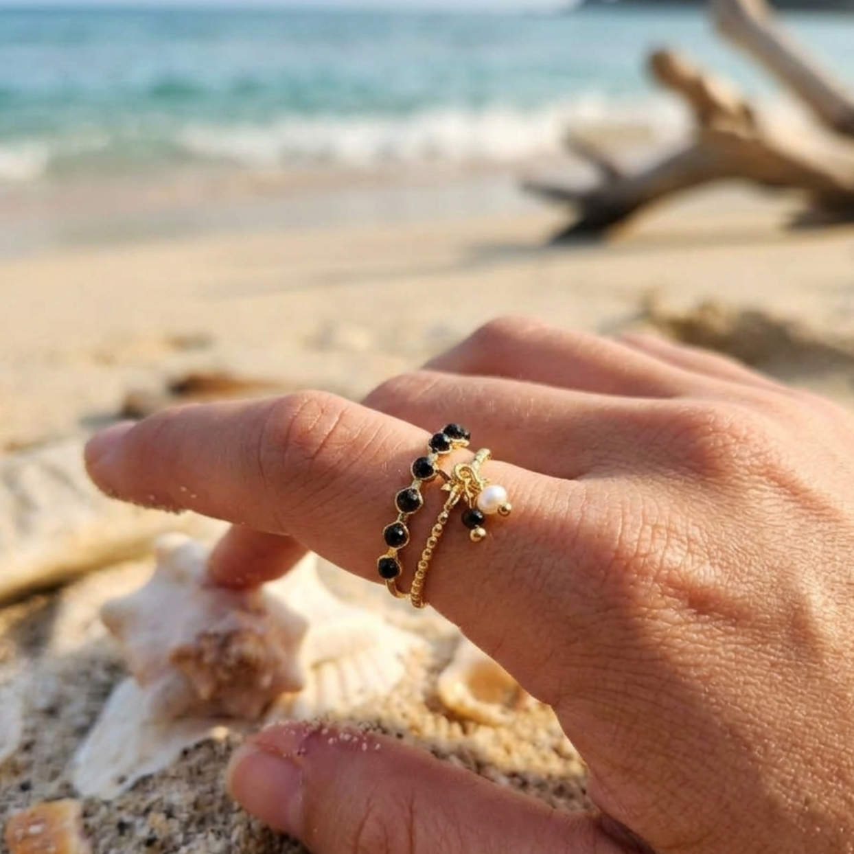 A close-up of a hand on a sandy beach wearing a delicate gold ring with black beads and a small pearl accent, with seashells and the ocean blurred in the background.