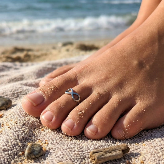 A silver infinity-shaped toe ring worn on a person's big toe, shown on a sandy beach with the ocean in the background.