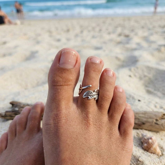 A silver toe ring with a leaf design worn on a person's big toe, photographed from a first-person perspective on a sandy beach with the ocean and distant beachgoers in the background.