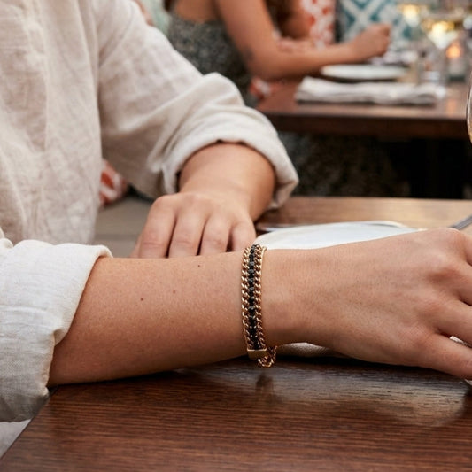 A close-up lifestyle shot of a woman at an outdoor evening dinner table wearing a gold chain bracelet. She is wearing a white linen shirt and holding a glass of white wine, with other diners and warm bokeh lights blurred in the background.