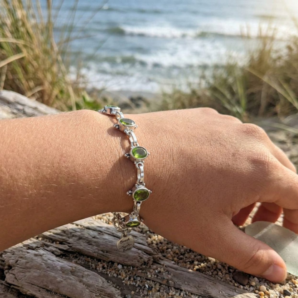 Hand wearing a silver bracelet with green gemstones on a beach.