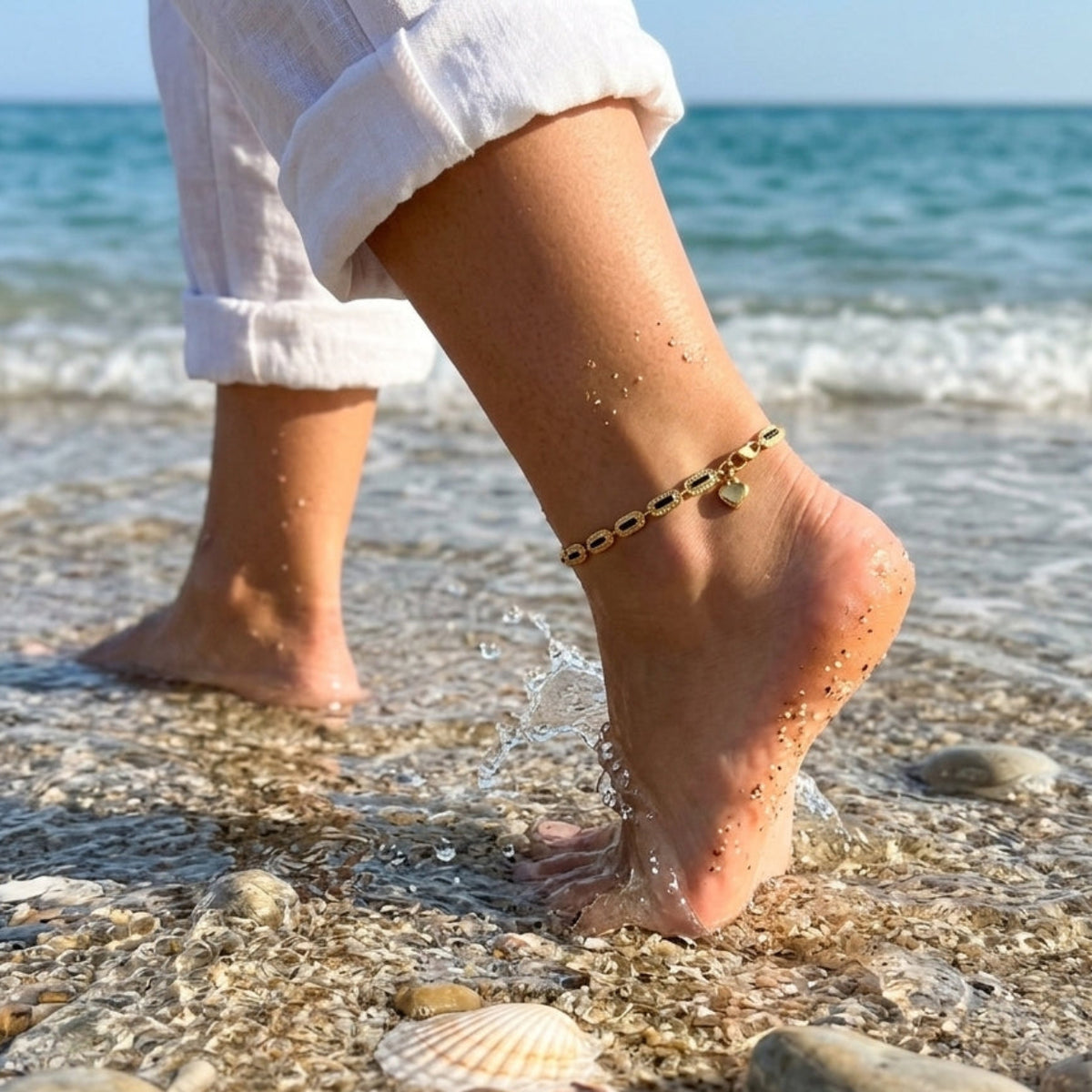A close-up lifestyle shot of a person walking on a sandy beach with ocean waves in the background, wearing a delicate gold anklet with small black and white beads on their ankle.