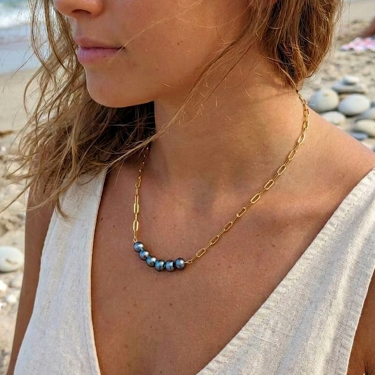 A woman on a sandy beach wearing a delicate gold chain necklace with a row of small light blue beads at the center.