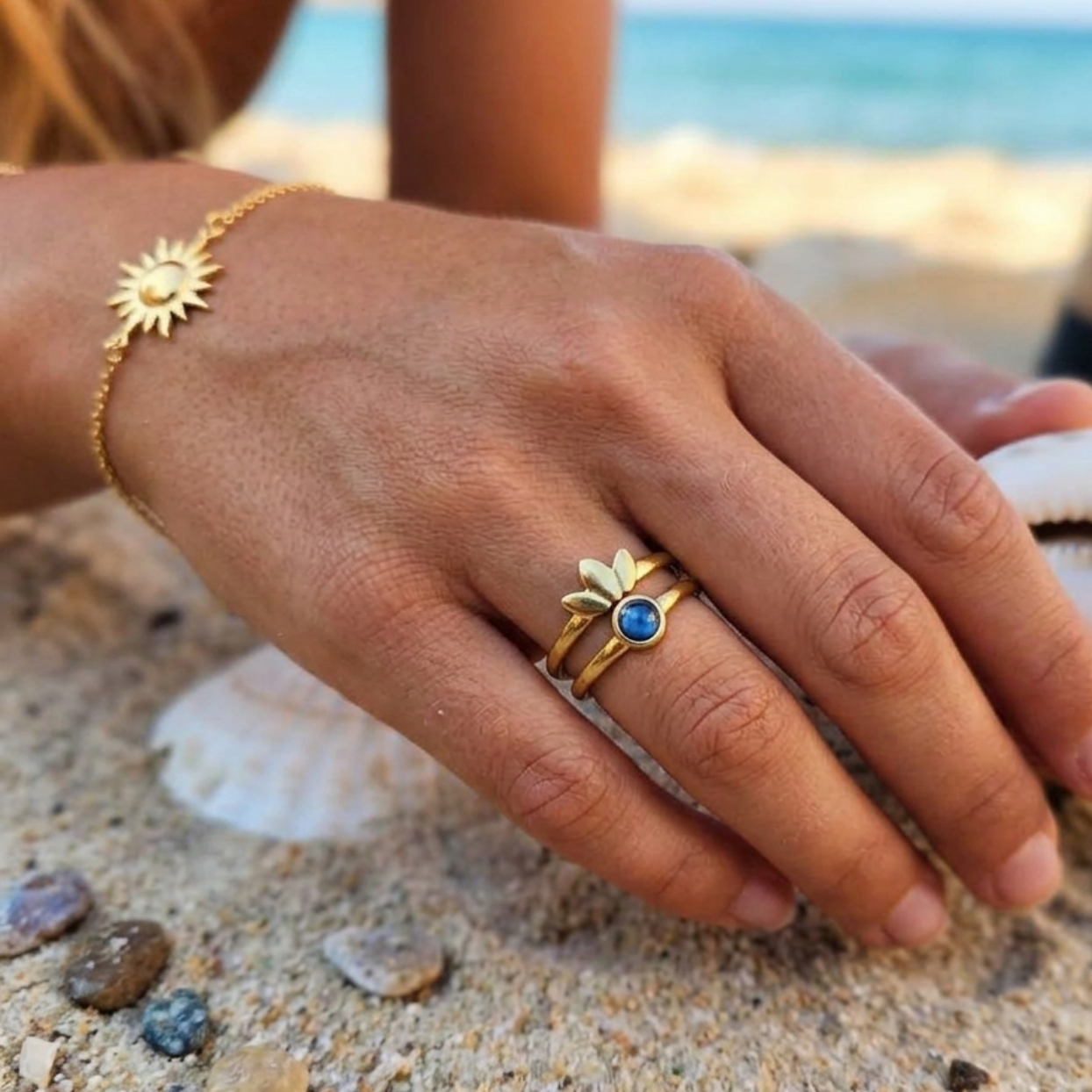 A close-up lifestyle shot of a person's hand on a sandy beach, wearing a gold ring with a blue oval stone and a gold sunburst bracelet.
