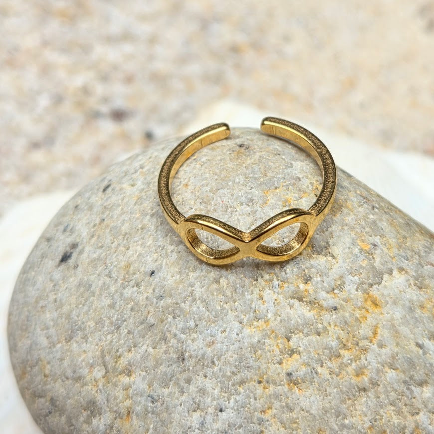 A gold open-ended ring with an infinity symbol design, resting on a smooth gray stone against a light beige textured background.