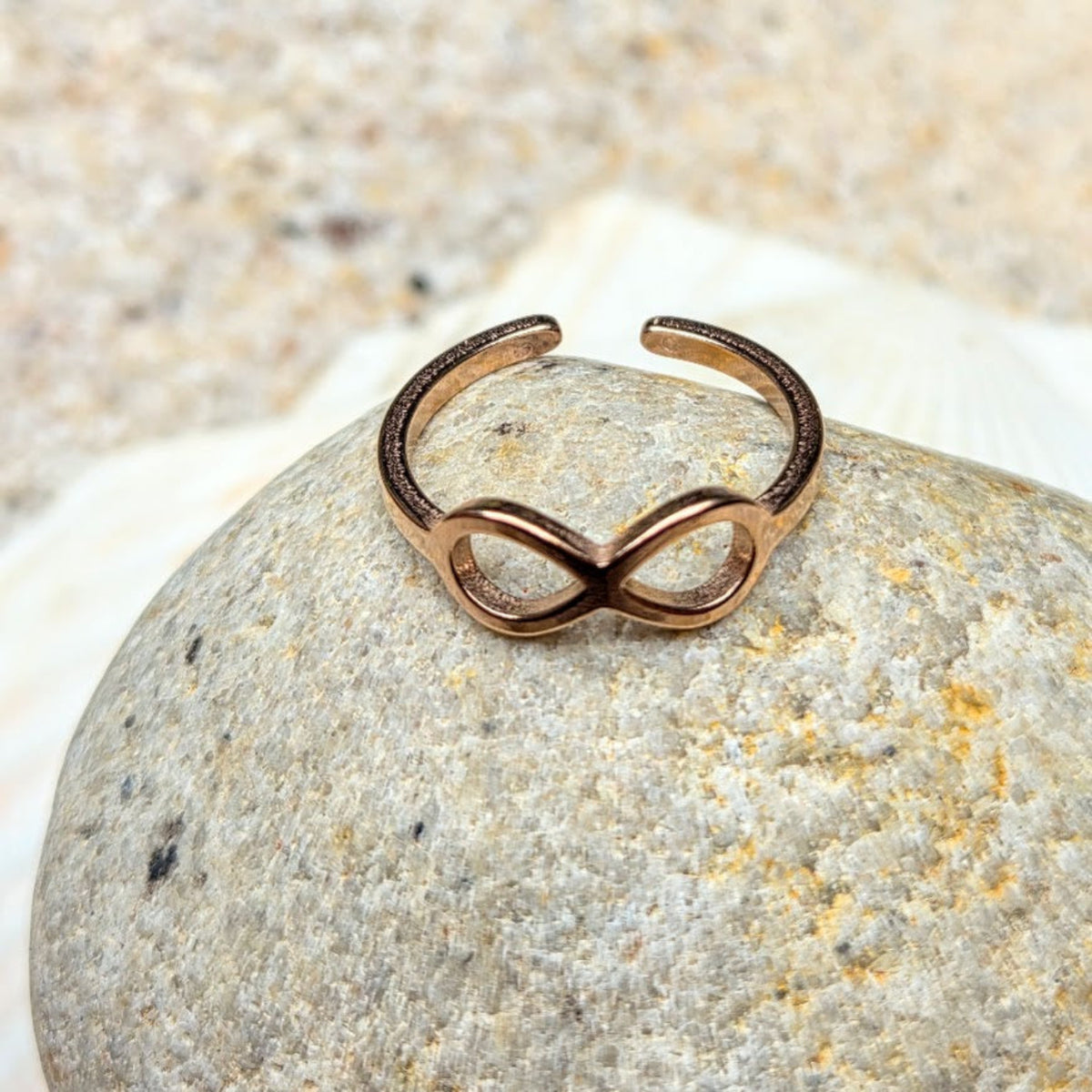 A rose gold infinity ring with a textured finish, displayed on a smooth gray stone with a white seashell in the background.