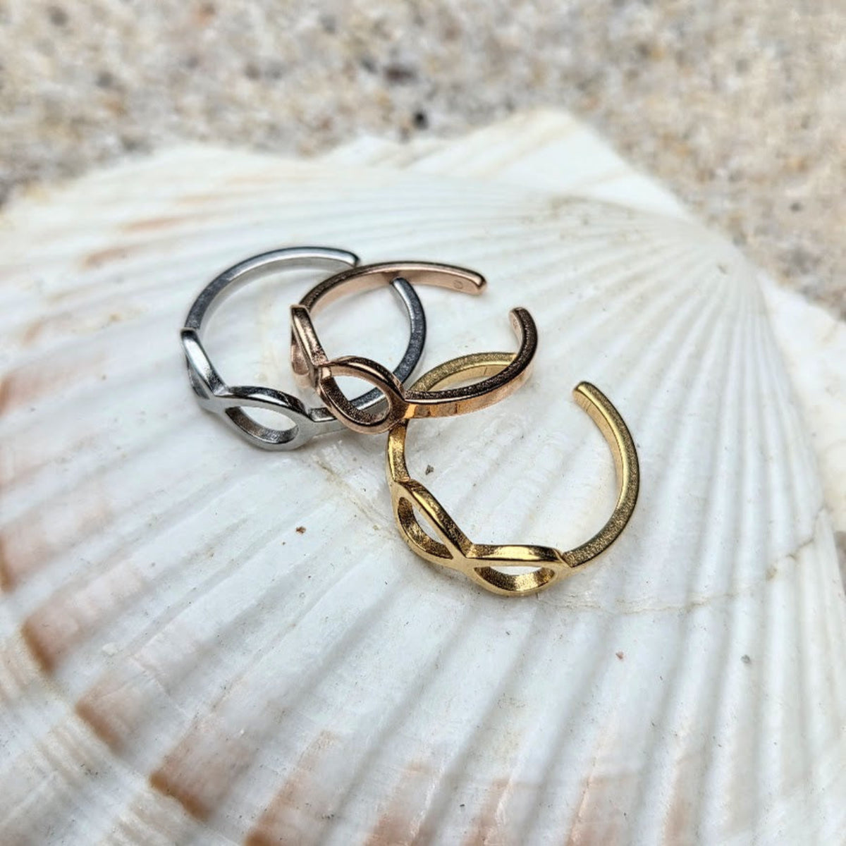 Three open infinity loop rings in silver, rose gold, and yellow gold, displayed on a large white seashell with sand in the background.