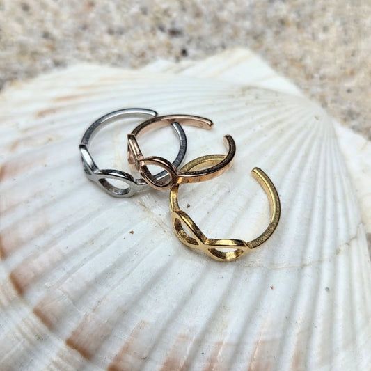 Three open infinity loop rings in silver, rose gold, and yellow gold, displayed on a large white seashell with sand in the background.