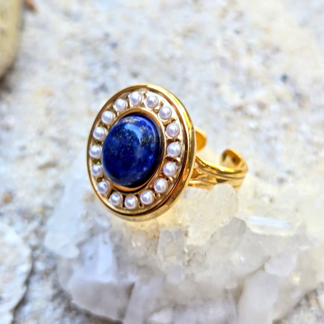 A gold ring featuring an oval blue lapis lazuli stone surrounded by a halo of small white pearls, displayed on a bed of white crystals.