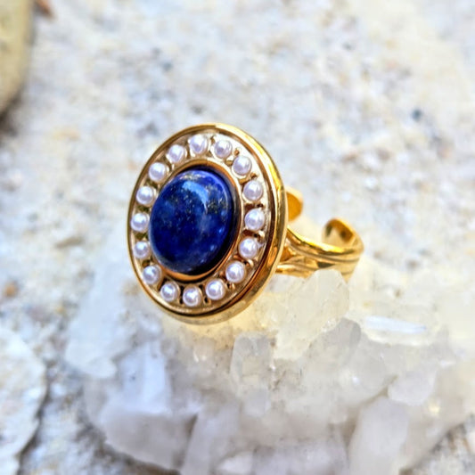 A gold ring featuring an oval blue lapis lazuli stone surrounded by a halo of small white pearls, displayed on a bed of white crystals.