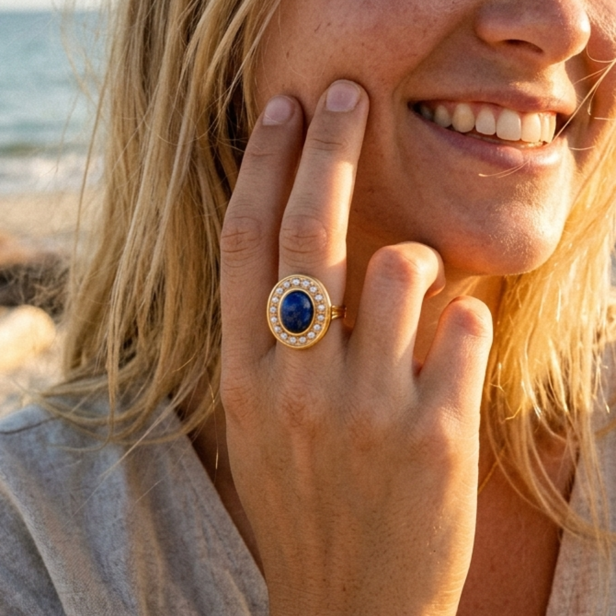 A smiling blonde woman on a beach wearing a gold ring with a large oval blue gemstone surrounded by small clear stones.