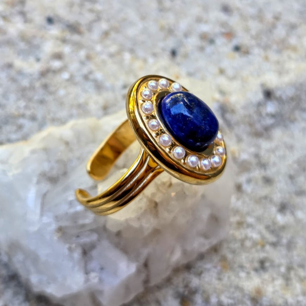 A gold ring with a deep blue oval center stone and a halo of small white pearls, displayed on a white crystal against a sandy, textured background.