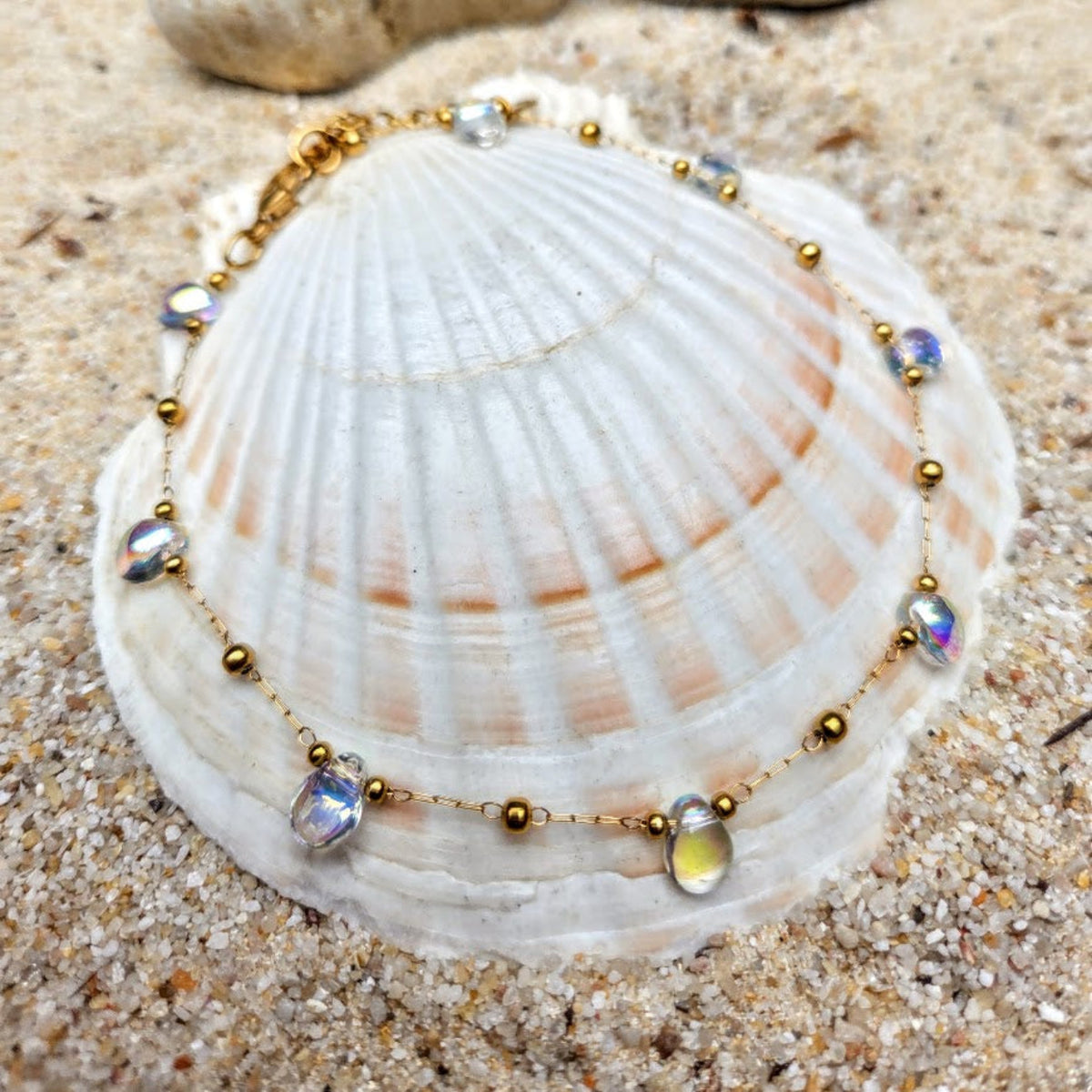A gold beaded necklace with iridescent shell-like beads is draped over a large white seashell resting on sandy ground.