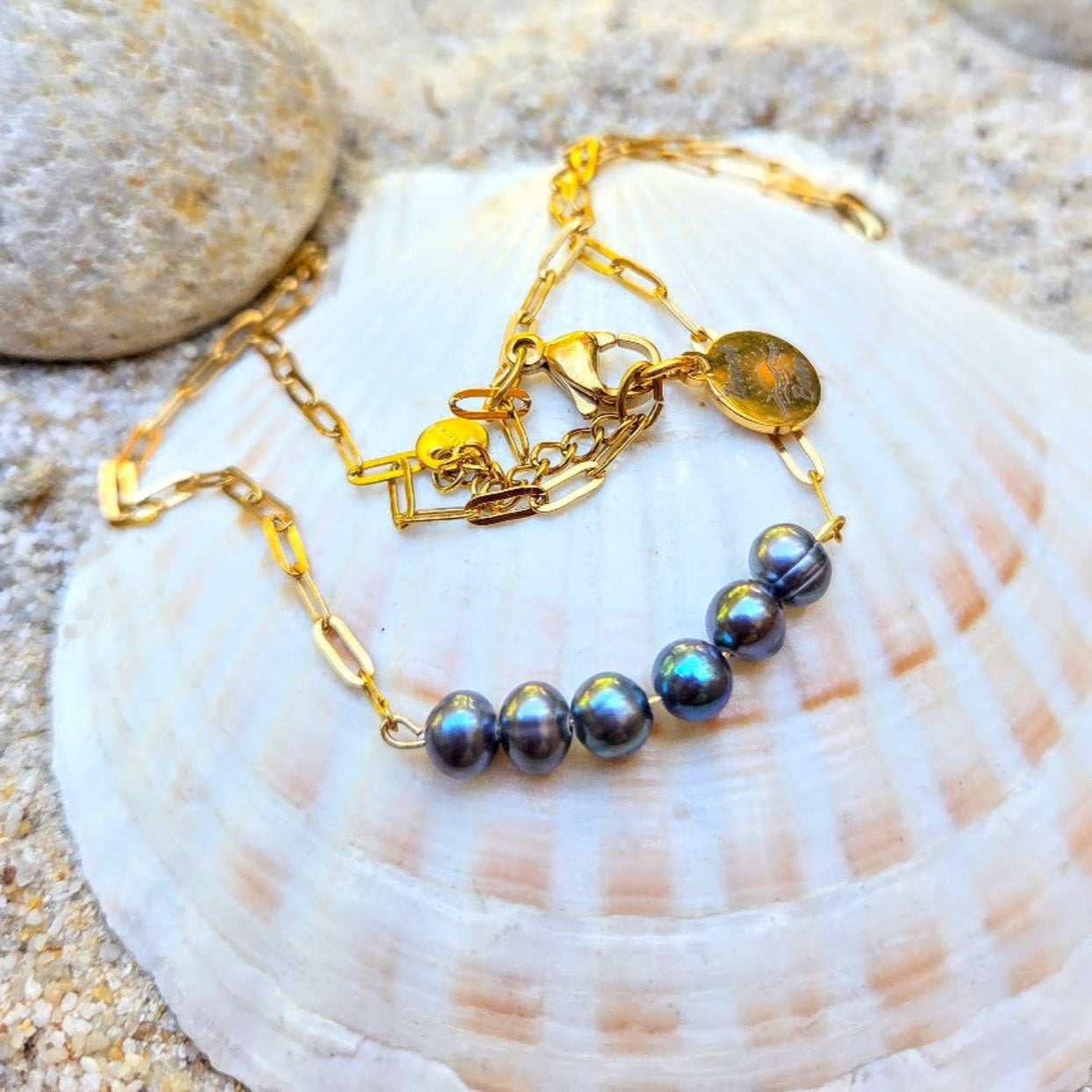 A gold chain necklace with five iridescent dark gray pearls, displayed on a large white seashell with sand and rocks in the background.