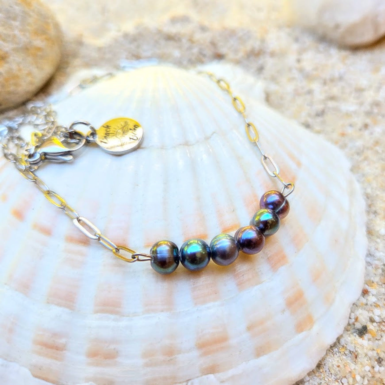 A silver chain bracelet with five iridescent dark pearl-like beads and a gold-tone charm, displayed on a large white seashell over sand.