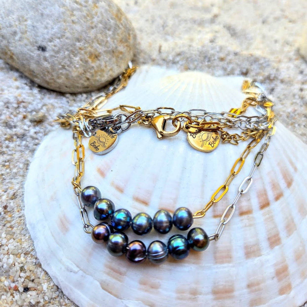 A multi-strand bracelet featuring iridescent dark pearls, gold and silver chains, and heart-shaped charms, displayed on a white seashell over sand.