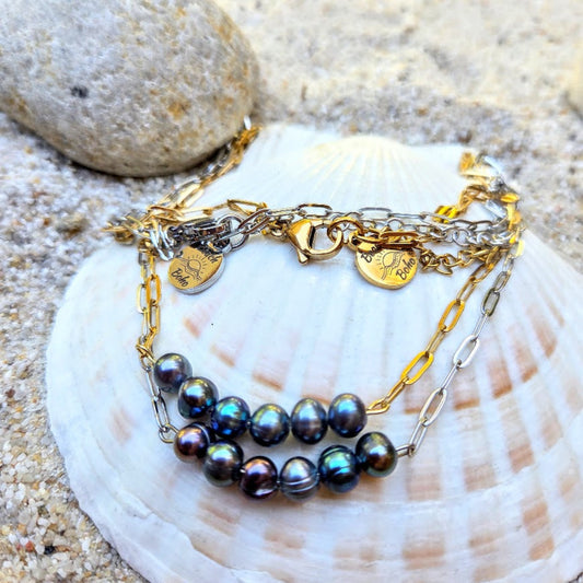 A multi-strand bracelet featuring iridescent dark pearls, gold and silver chains, and heart-shaped charms, displayed on a white seashell over sand.