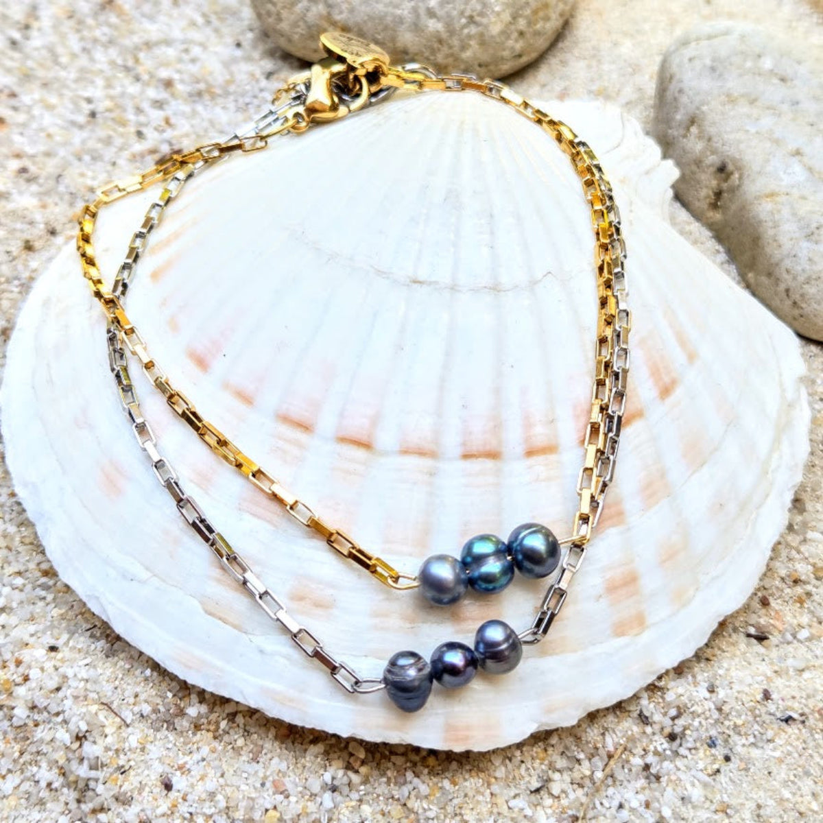 A double-strand necklace featuring one gold chain and one silver chain, both adorned with dark gray baroque pearls, displayed on a white seashell over sand.