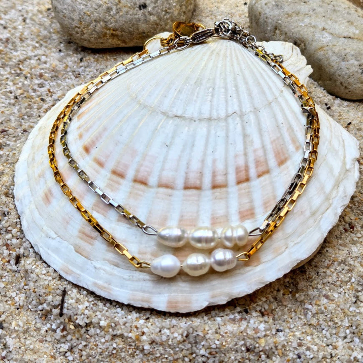 A double-strand bracelet featuring a gold and silver chain with three white baroque pearls, displayed on a large white seashell over sand.