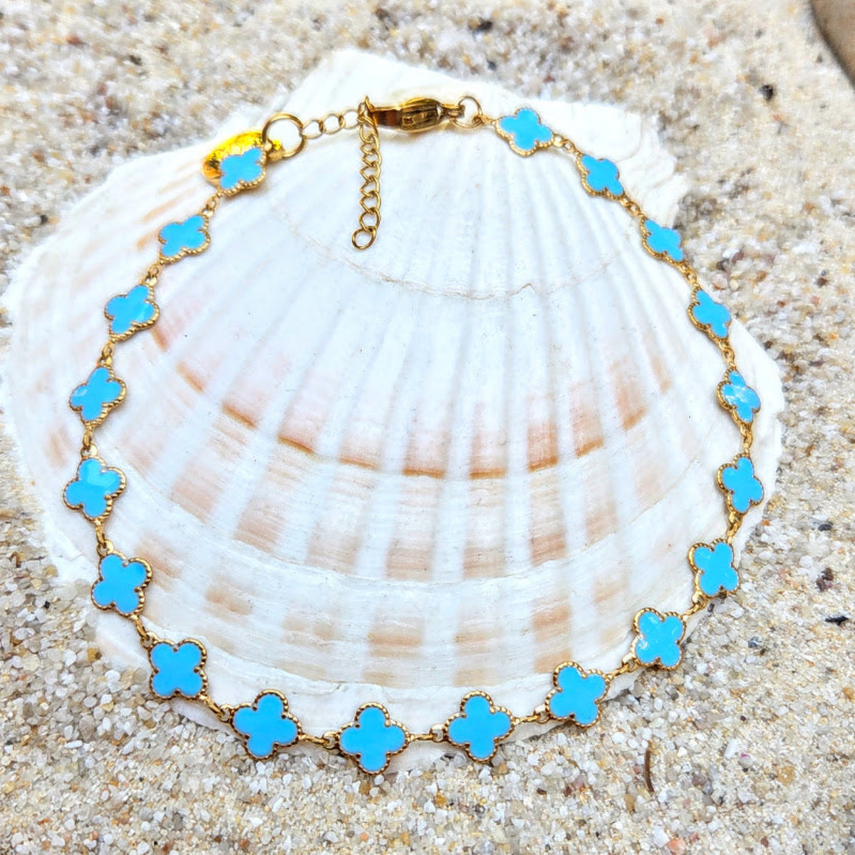 A gold chain bracelet with turquoise blue four-petal clover motifs, displayed on a large white seashell over sandy ground.