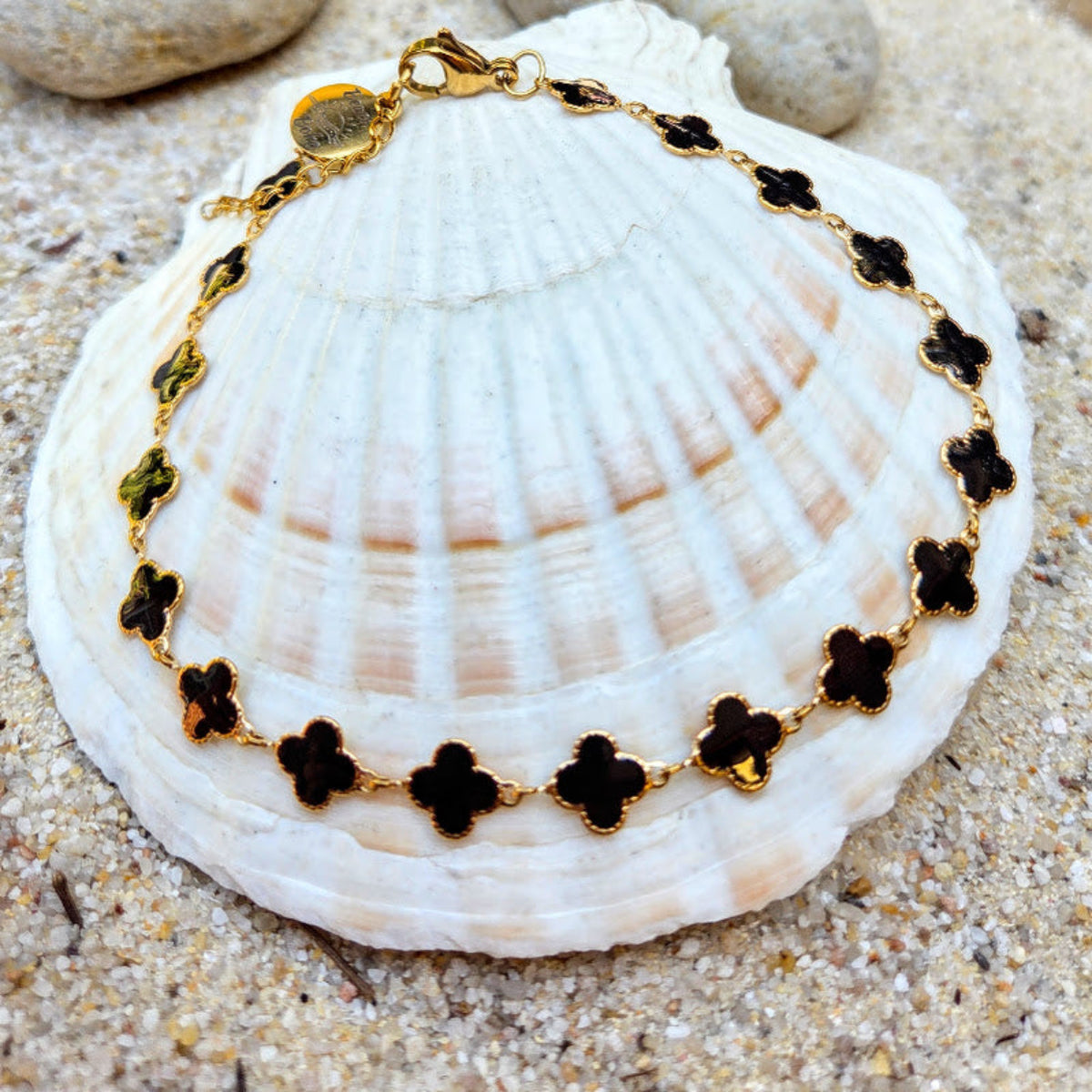 A gold chain bracelet with black clover-shaped charms, displayed on a large white seashell over sand.