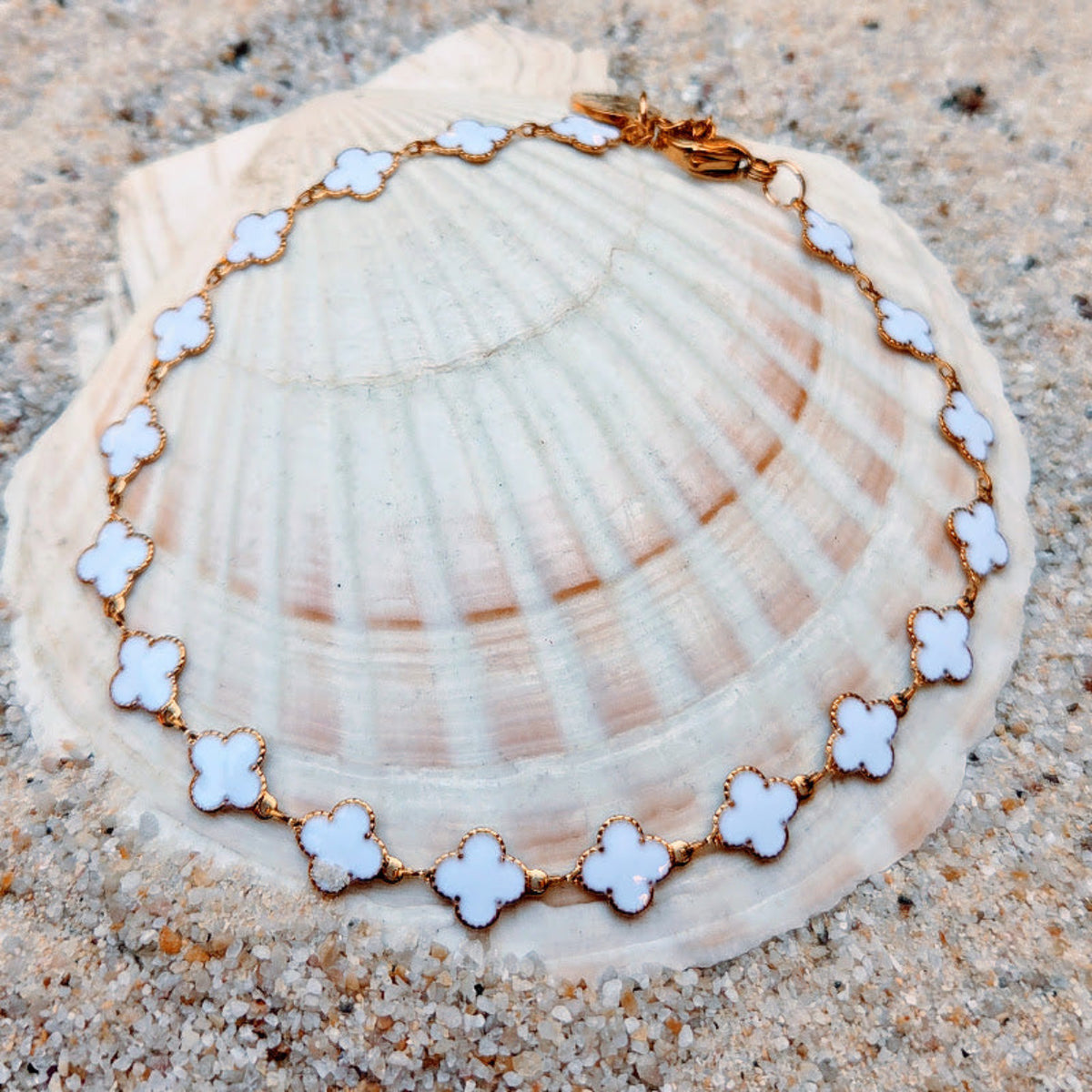 A gold chain bracelet with white enamel clover motifs, displayed on a large seashell over sand.