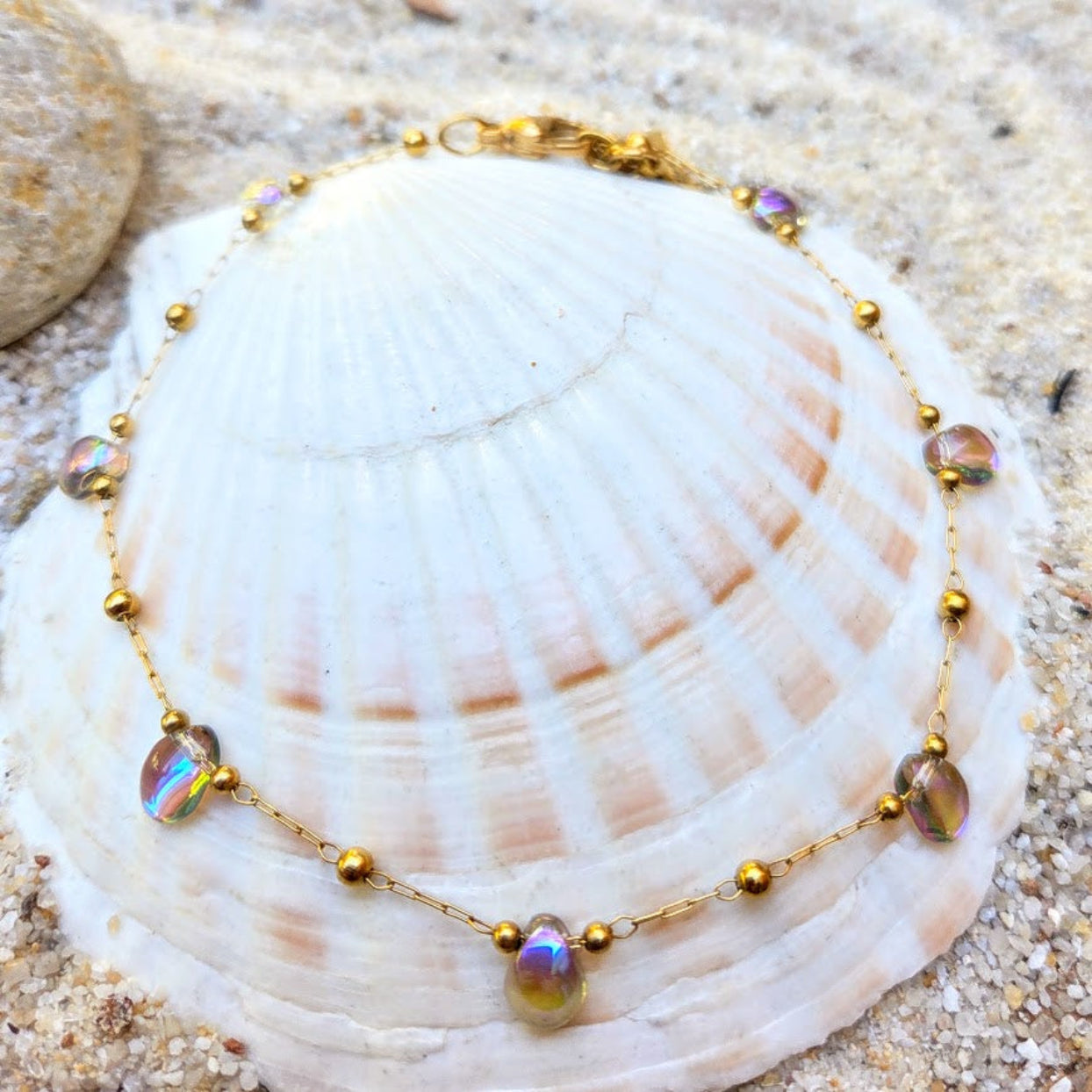 A gold beaded chain bracelet with iridescent oval beads, displayed on a large white seashell over sand.