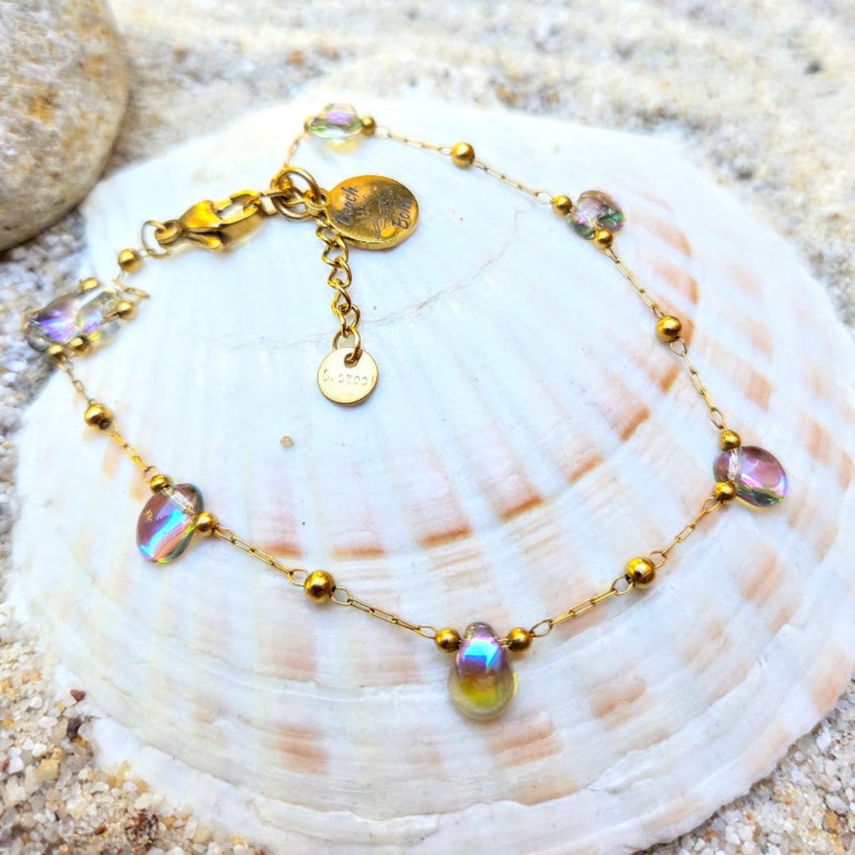 A gold chain bracelet with iridescent teardrop beads and small gold beads, displayed on a large white seashell over sand.