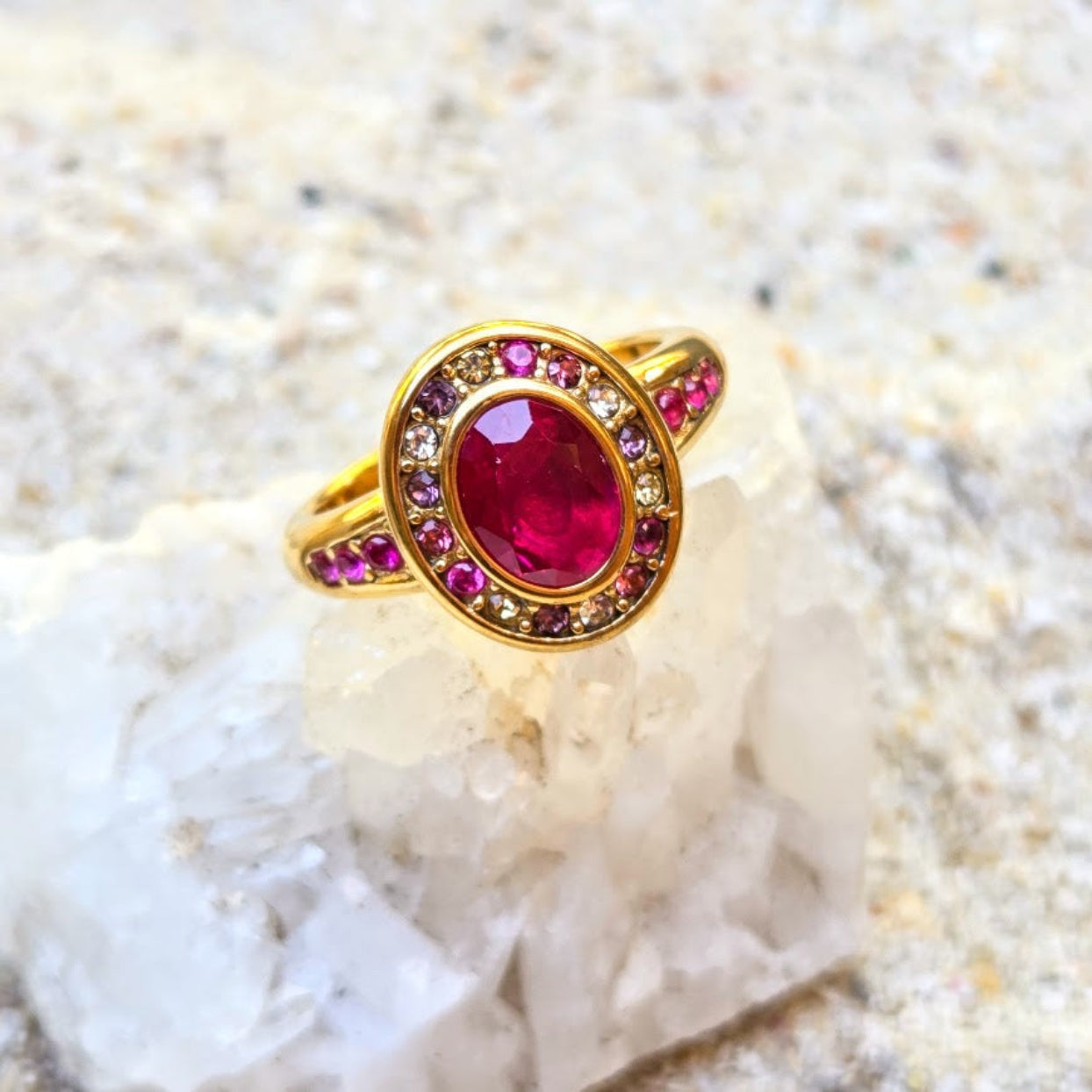 A gold ring with a large oval pinkish-red center stone and smaller pink and clear stones around it, displayed on a white crystal against a sandy beige background.